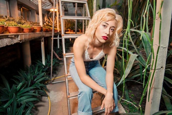 Beautiful female model with short curly hair wearing jeans and top with bear shoulders sitting on a stepladder surrounded by succulents. Looking at camera at botanical garden.