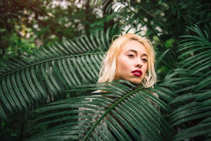 Closeup portrait of sensual female fashion model with damp blond hair and red lips standing behind the palm branch, covered in it. Having a dreamy look, at the botanical garden.
